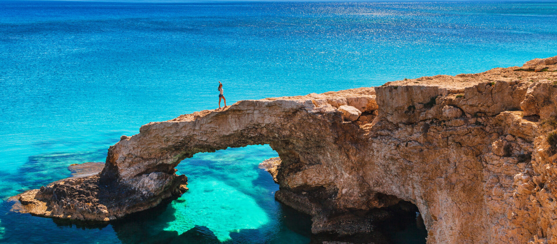 Woman on the beautiful natural rock arch near of Ayia Napa, Cavo Greco and Protaras on Cyprus island, Mediterranean Sea. Legendary bridge lovers. Amazing blue green sea and sunny day.